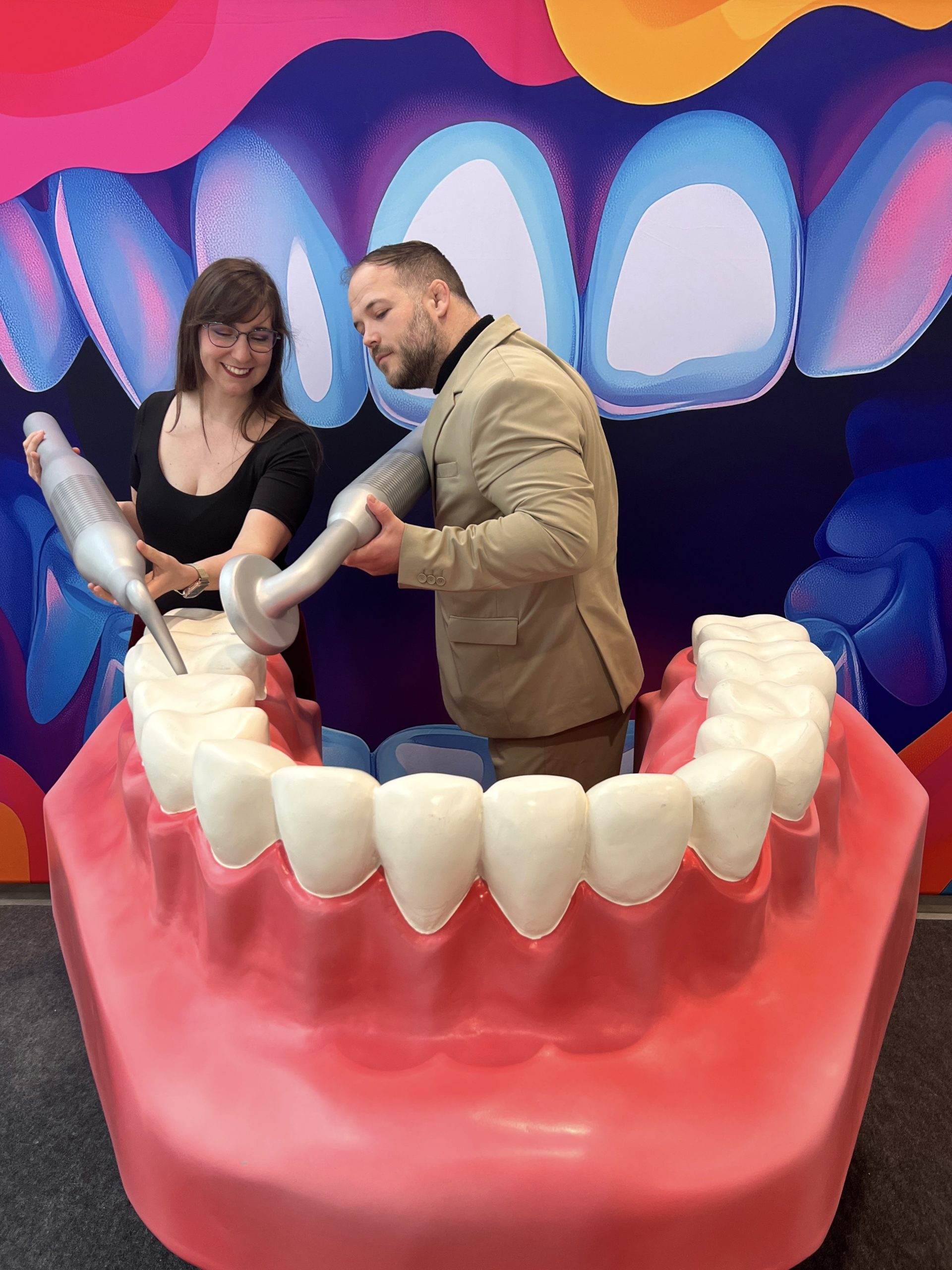 A képen egy látványos fogászati kiállításon készült interaktív bemutató látható, ahol szakemberek óriási fogmodellen szemléltetik a professzionális fogászati kezeléseket. Az innovatív eszközök és a lenyűgöző vizuális háttér tökéletesen tükrözik a digitális fogászat fejlődését és a legmodernebb technológiák alkalmazását.